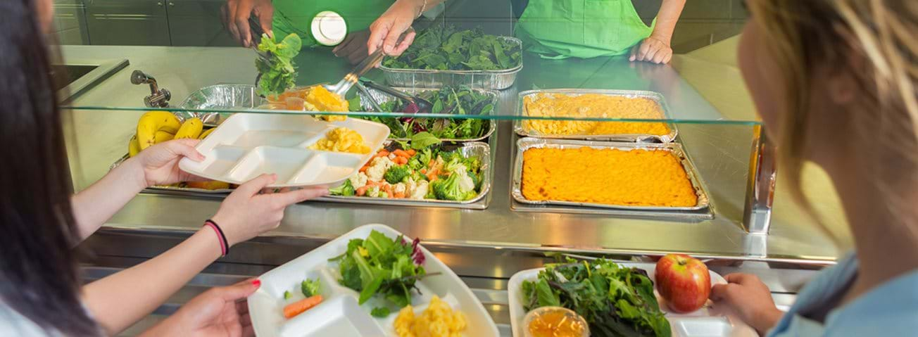 Children line up with trays while being served at a school lunch counter.