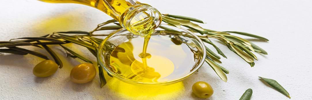 A bottle of olive oil filling a glass bowl on a counter with fresh olives